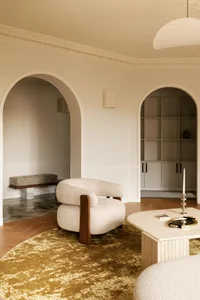 Modern living room featuring a curved cream linen armchair with walnut arms, a large travertine coffee table, mustard-yellow abstract rug, and multiple arched doorways leading to built-in shelving.