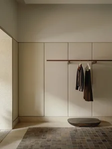 Minimalist entryway with beige terrazzo wall, built-in cabinetry, wooden clothing rack holding a leather jacket and brown dress, and a dark live-edge wooden bench on a brown tiled floor.