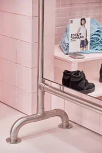 Close-up of a stainless steel pipe mirror mount and railing in a retail store with pink square tiles and black sneakers displayed on a shelf.