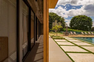 View from a modern house interior looking out onto a travertine stone pool terrace with wooden ceiling overhang, lounge chairs, and lush garden under a blue sky.