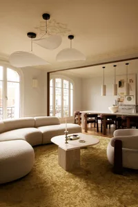 Spacious Parisian living and dining room featuring a large curved white boucle sofa, irregular marble coffee table, mustard-yellow rug, and three large white mushroom-shaped pendant lights hanging from a high ceiling with ornate molding.