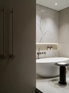 Modern minimalist bathroom featuring a white freestanding oval bathtub, large marble tile walls, recessed lighting, and a black cylindrical side table with folded white towels.
