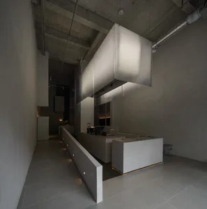 Modern brutalist commercial kitchen interior featuring a long concrete service counter, central kitchen island, and large rectangular fabric pendant lights suspended from an exposed concrete ceiling.