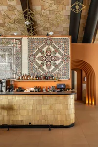 Modern bar counter with light wood shingle cladding, terracotta orange walls, intricate colorful patterned tiles, and hanging geometric brass pendant lights.