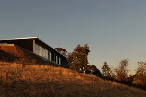 Modern single-story concrete and glass house built into a dry grass hillside during warm golden hour sunset, surrounded by oak trees under a clear blue sky.