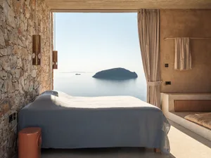 Minimalist bedroom with rough stone wall, concrete elements, large bed with grey linen, and a wide opening revealing a calm sea and distant island under bright daylight.