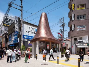Pedestrians walking past a distinctive copper conical building with a pointed roof on a busy street corner in Tokyo, surrounded by shops and overhead utility wires under a clear blue sky.