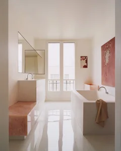 Minimalist bathroom interior with white walls, glossy floor, pink terrazzo bench and bathtub, large mirror, and natural light from glass doors.