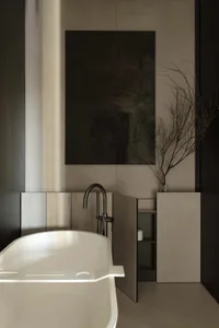 Modern minimalist bathroom featuring a white freestanding bathtub, matte concrete walls, black matte faucet, dark abstract painting, and dried branches in a glass vase.