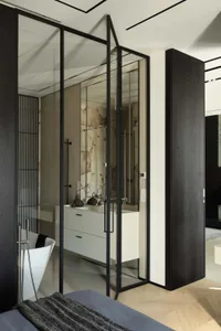 Modern ensuite bathroom viewed from bedroom, featuring black steel and glass folding partition, white floating vanity, marble accent wall, and freestanding white bathtub.