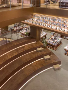 View from above of a curved wooden staircase with integrated LED lighting leading to the second floor of a spacious, modern bookstore with extensive wooden bookshelves and display tables.