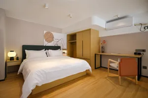 Modern minimalist bedroom with dark green upholstered bed, white linens, light oak wardrobe and desk, and an orange leather chair under warm lighting.