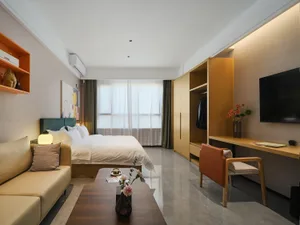 Modern hotel bedroom with teal velvet headboard, oak wardrobe and desk, beige sofa, and large window with sheer curtains.
