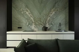 Modern open-plan kitchen and living area featuring a large sage green marble feature wall, white minimalist cabinetry, black fixtures, and a dark green sofa in the foreground.