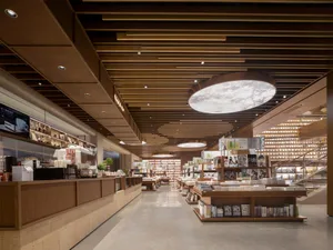 Interior view of a modern bookstore café with warm wooden slat ceilings, a large circular skylight fixture, built-in bookshelves, and a service counter on the left.