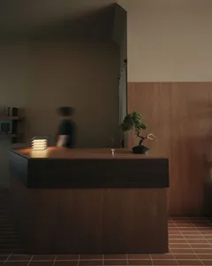 Modern minimalist reception desk made of dark walnut wood with a lit table lamp, burning incense, and a bonsai tree against a warm wooden wall panel.