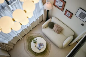 Top-down view of a modern living room with a curved cream boucle sofa, two decorative pillows, a round white coffee table on a layered green and beige organic-shaped rug, a cloud-like multi-sphere pendant light, and framed abstract art on the wall.