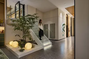 Modern hotel lobby with white floating staircase, glass railing, large indoor tree, three glowing spherical moon lamps on a white pebble platform, and a green neon logo on the wall.