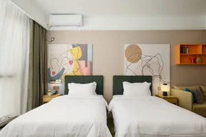 Modern hotel twin room with two beds dressed in white linens, dark green velvet headboards, colorful abstract wall art, wooden nightstands, and an orange wall shelf.