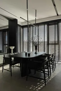 Modern minimalist dining room featuring a black oak rectangular table surrounded by six black leather chairs, dramatic linear pendant lights, large windows with horizontal blinds, and a tall black storage unit.