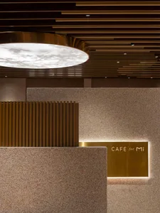 Modern cafe counter featuring speckled terrazzo walls, vertical wood slats, a large circular backlit ceiling fixture, and an illuminated brass sign reading 'CAFE for Mi'.