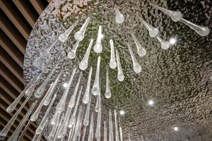 Close-up view looking up at an artistic chandelier featuring numerous clear glass teardrop and rod elements hanging from a large circular hammered silver ceiling panel with integrated spotlights.