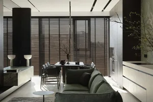 Modern minimalist open-plan dining and living room with sage green sofa, white marble dining table, dark wooden blinds, and integrated cabinetry bathed in natural sunlight.