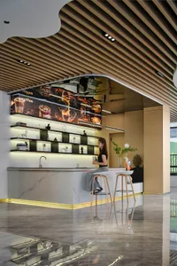 Woman sitting at a modern coffee bar with white marble counter, backlit display shelves, and wooden slat ceiling in a commercial space.