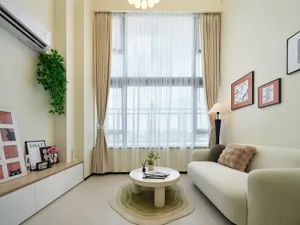 Modern minimalist living room with a sage green boucle sofa, curved white coffee table on a green rug, large window with sheer curtains, and built-in low cabinet displaying art and plants.