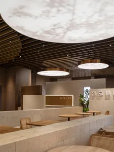 Modern café interior featuring light-colored terrazzo counters, wooden tables, rattan chairs, and large circular pendant lights with leaf-patterned projections on the curved wooden slat ceiling.