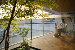Modern cafe veranda featuring warm wood deck flooring, large glass windows, an indoor tree, and a woman sitting at a round table enjoying coffee in natural sunlight.
