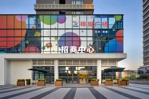 Front view of Zhangqiao Plaza commercial building with large glass facade covered in colorful bubble and gift mural, outdoor seating with green umbrellas and planter boxes in the foreground.