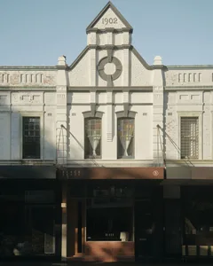 一座兩層樓白色粉刷的1902年歷史建築正面，擁有裝飾性山牆、圓形圖騰、懸掛窗簾的木框窗戶，以及Black Blaze店面的現代棕色遮陽棚。