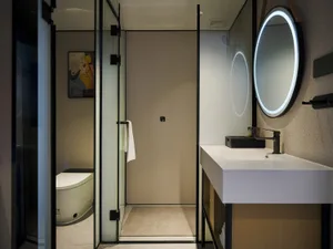 Modern bathroom interior with white floating vanity, black matte faucet, large circular backlit mirror, terrazzo walls, and glass-enclosed shower area with toilet visible in background.