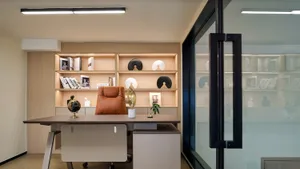 Modern minimalist home office with a cognac leather executive chair, wooden desk, backlit light-wood shelving displaying books and decorative objects, and a glass partition wall.