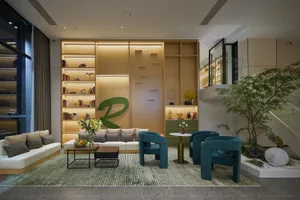 Modern living room with white sofa, teal velvet armchairs, built-in illuminated wooden shelves, green accent wall art, and an indoor garden area with a tree under a glass staircase.