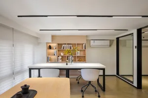 Modern minimalist home office with white rectangular table, white ergonomic chairs, light oak built-in bookshelf, and black linear ceiling lights.