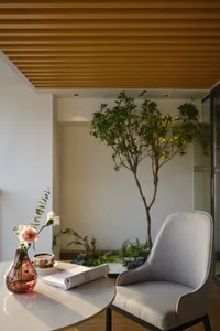 Modern reading nook featuring a grey upholstered armchair, round white marble table with pink flower vase and open book, set against a lush indoor tree and warm wooden slat ceiling.