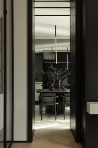 View from a hallway through a dark wood door frame into a modern minimalist dining room with dark table, leather chairs, pendant lights, and a large sculptural branch arrangement in a vase.