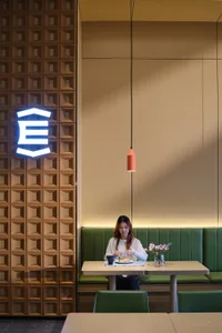 Woman dining alone at a light wood table in a modern restaurant booth with green upholstery, a coral pendant lamp, and a striking textured wooden wall featuring a glowing neon logo.