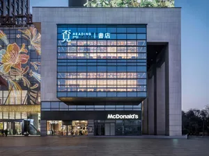 Night view of a modern commercial building featuring a glowing double-height glass reading room with bookshelves and the Reading Mi sign, positioned above a McDonald’s restaurant with festive floral light installations on the adjacent structure.