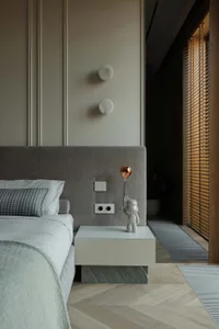 Modern minimalist bedroom featuring a grey upholstered headboard, white and marble bedside table with a copper balloon dog lamp, herringbone wood flooring, and wooden window blinds.