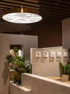Modern restaurant interior featuring textured stone partitions, large round brass pendant light, wooden dining table, rattan chair and lush green plants