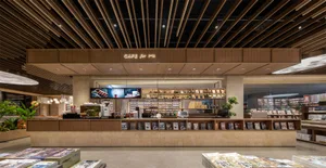 Modern bookstore cafe interior featuring a long travertine and wood service counter, linear wood slat ceiling, warm neutral palette, and integrated book displays.