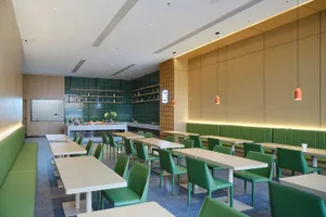 Modern cafeteria interior with rows of white tables, green leather chairs and booths, green tiled feature wall, and warm wood paneling under pendant lighting.