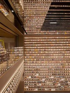 Multi-level interior of a modern bookstore featuring an enormous floor-to-ceiling wooden bookshelf wall filled with colorful books, dramatic mirrored ceiling reflecting the shelves into infinity, warm ambient lighting, and glass railing overlooking lower levels.