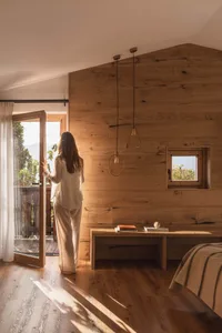 Woman in cream outfit standing by open glass door in a modern wooden bedroom with oak walls, pendant lights, and mountain view.