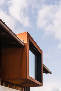 Low-angle view of a rust-colored corten steel cantilevered window box extending from a building roofline with blue sky and clouds in the background.