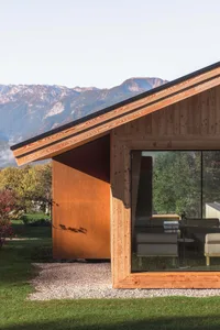 Exterior view of a modern wooden cabin featuring a large Corten steel panel, expansive glass windows revealing the living area, set against a scenic mountain range with morning mist.