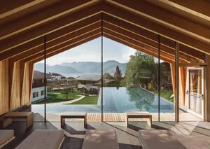 Modern wooden villa interior featuring an indoor swimming pool, minimalist stone and wood furniture, and panoramic mountain views through large glass walls under a pitched timber ceiling.
